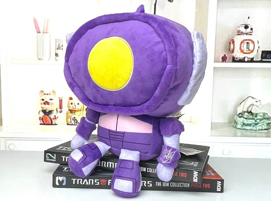 Purple and yellow plush toy on top of a book with shelves in the background