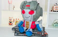 Load image into Gallery viewer, Plush toy resembling a robot character on top of Transformers books with a white shelf in the background.