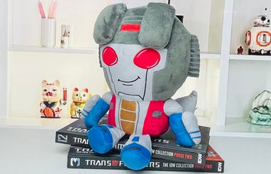 Load image into Gallery viewer, Plush toy resembling a robot character on top of Transformers books with a white shelf in the background.