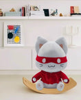 Load image into Gallery viewer, Plush toy cat in a red outfit on a wooden stand with a white shelf in the background