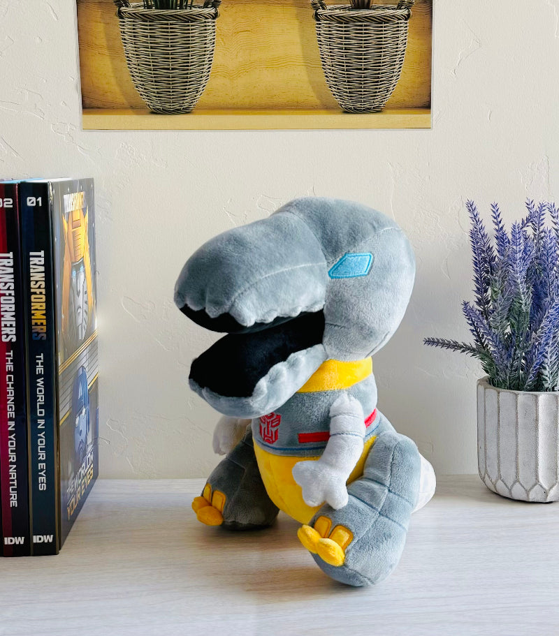Plush toy resembling a robot dinosaur on a shelf with books and a plant in the background