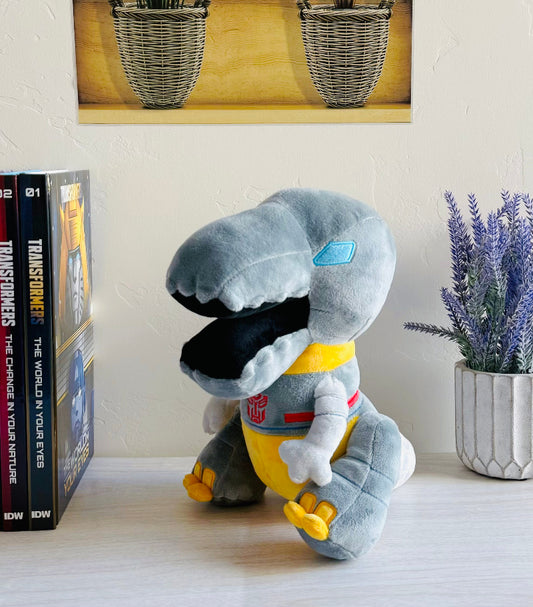 Plush toy resembling a robot dinosaur on a shelf with books and a plant in the background