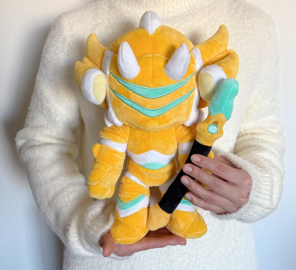 Plush toy resembling a dragon with yellow, white, and teal colors held by a person wearing a white sweater.