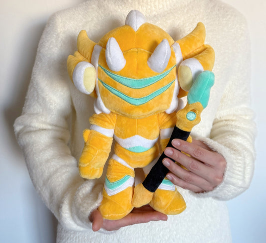 Plush toy resembling a dragon with yellow, white, and teal colors held by a person wearing a white sweater.