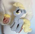 Load image into Gallery viewer, Plush toy of a gray pony with yellow hair held by a person wearing a white fleece jacket.