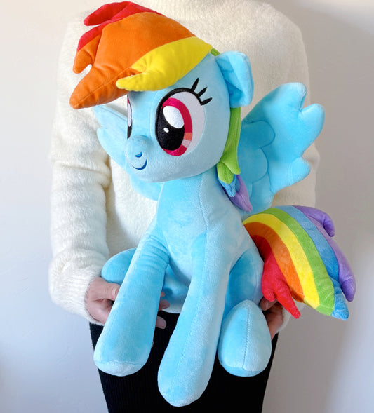 Plush toy of a blue pony with rainbow hair and wings held by a person against a white background