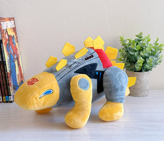 Plush toy resembling a robot dinosaur on a table with books and a plant in the background