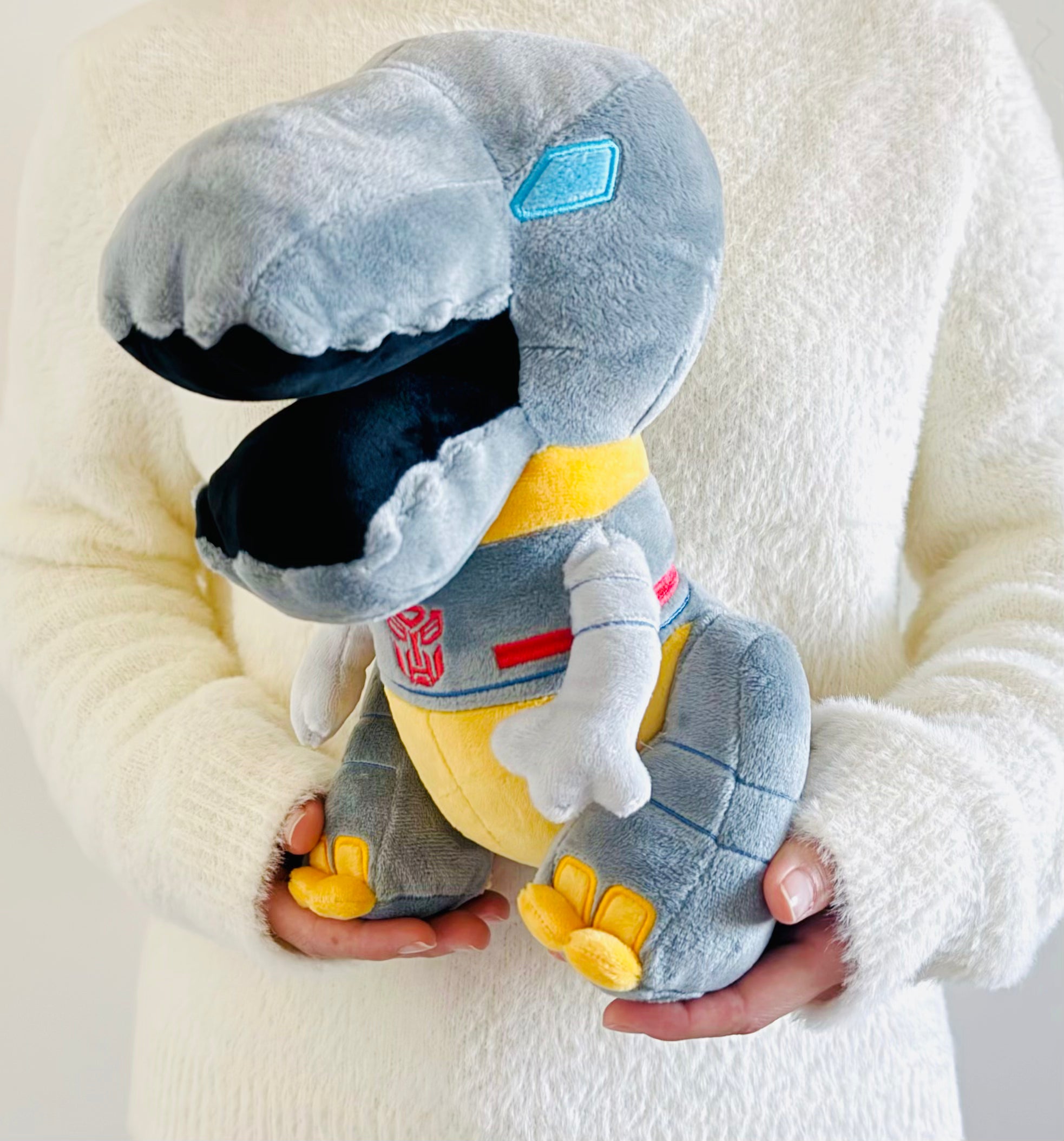 Plush toy resembling a robot dinosaur held by a person wearing a white sweater.
