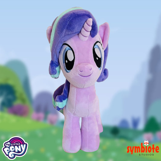 Plush toy of a purple pony with a green mane against a blurred outdoor background, featuring 'My Little Pony' and 'symbiote STUDIOS' logos.