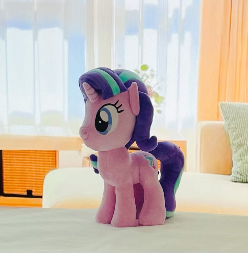 Plush toy resembling a pony with a colorful mane on a bed in a bright room.