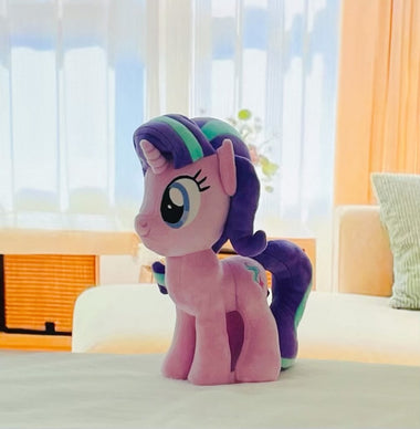 Load image into Gallery viewer, Plush toy resembling a pony with a colorful mane on a bed in a bright room.

